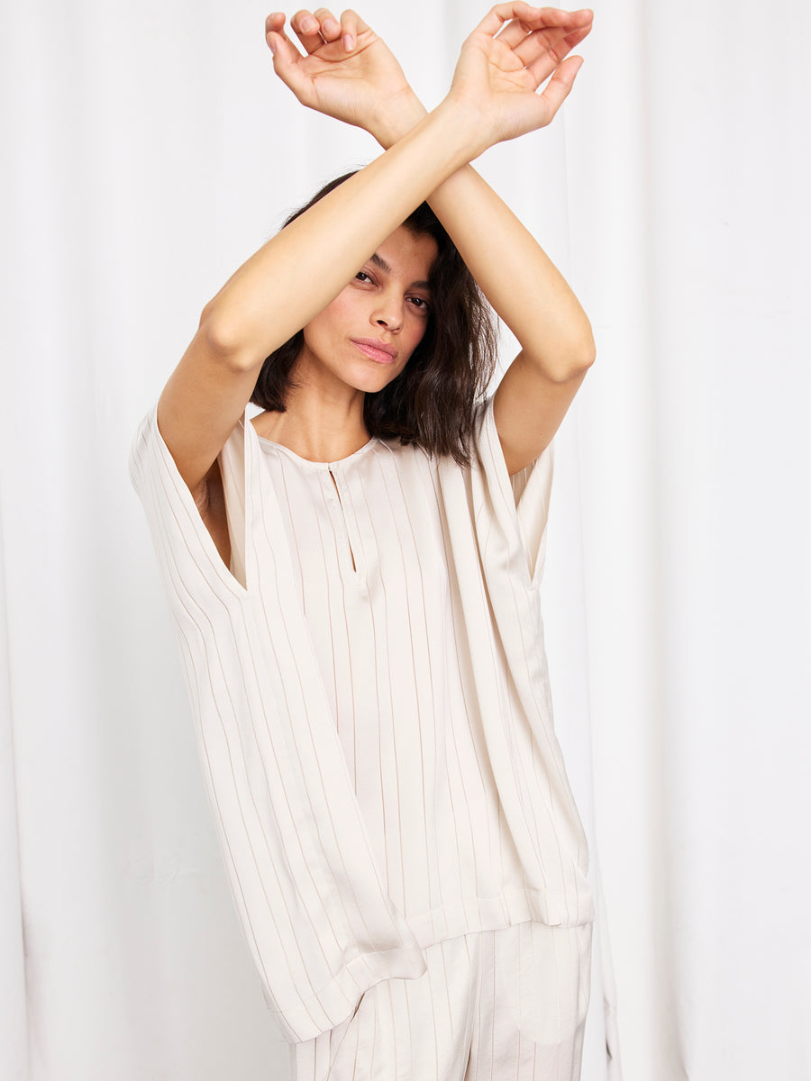 A woman wearing the Rae Poncho Top by Zero + Maria Cornejo stands with her arms crossed above her head against a white curtain background.