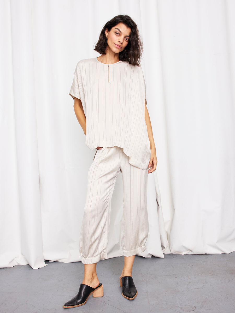 A woman with short, dark hair stands before a white curtain, wearing the Rae Poncho Top and matching pants by Zero + Maria Cornejo, both made in New York, paired with black heeled mules.