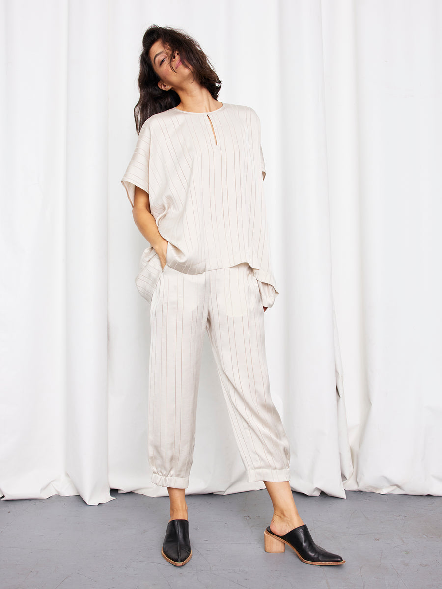 A woman models the Rae Poncho Top by Zero + Maria Cornejo—a loose, cream, pinstriped top with short sleeves—paired with cropped pants and black heeled mules. She stands with hands in her pockets against a plain white curtain backdrop.
