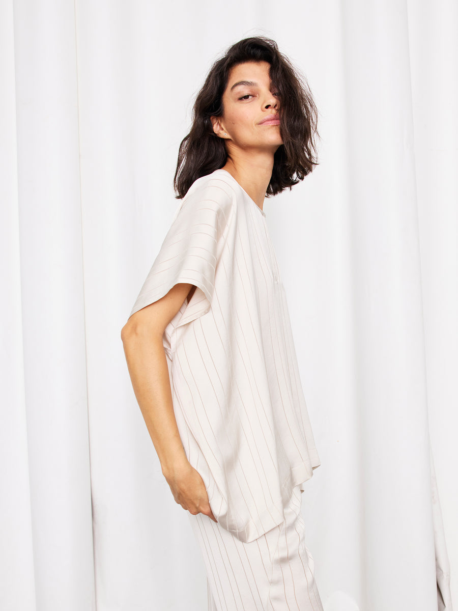 A person with shoulder-length dark hair poses sideways, wearing the Rae Poncho Top by Zero + Maria Cornejo— a loose, woven striped top with short sleeves—set against a white curtain backdrop.