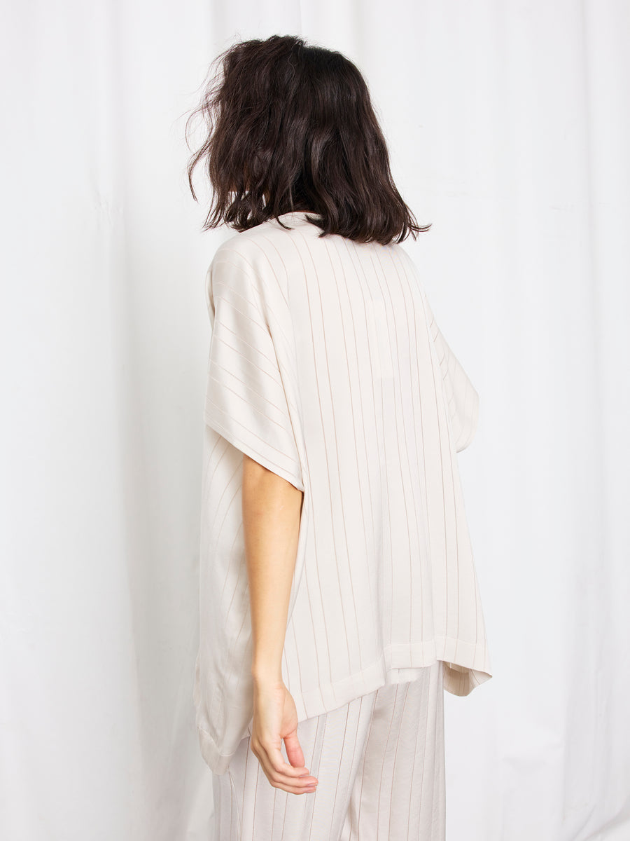 A person with shoulder-length dark hair stands facing away, wearing the Rae Poncho Top by Zero + Maria Cornejo and matching pants with subtle vertical stripes, set against a plain white background.