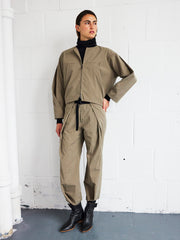 A person stands against a white wall wearing the Zero + Maria Cornejo Wrap Akeo Pant in khaki, a matching zip-up jacket, a black turtleneck, black boots, and is looking to the side.