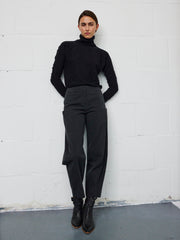 A person stands against a white brick wall, wearing a black turtleneck and the Seamed Margot Pant by Zero + Maria Cornejo—wide-leg cropped pants crafted from cotton blend twill. With their hands in their pockets and gaze fixed on the camera, they embody timeless style made in New York.