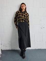 A woman stands against a white brick wall wearing a yellow and black plaid shirt, the Ido Skirt by Zero + Maria Cornejo—a woven mid-length skirt with an asymmetrical hem—and black ankle boots.