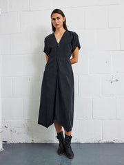 A person stands against a white brick wall, wearing the Aissa Joi Dress by Zero + Maria Cornejo—a black, cotton blend V-neck midi dress with short sleeves and an asymmetrical hem—paired with black ankle boots. Their hands are behind their back and they have a neutral expression.