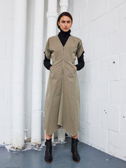 A person stands indoors against a white brick wall, wearing the Zero + Maria Cornejo Aissa Joi Dress with an A-line skirt over a black turtleneck, paired with black ankle boots. Their hands are in their pockets and they look directly at the camera.