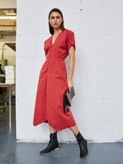 Wearing the Aissa Joi Dress by Zero + Maria Cornejo—a red, cotton blend, mid-length dress with an A-line skirt—a person stands against a white wall in what looks like an industrial or studio space. They pair the dress with black ankle boots and hold a brown clutch.