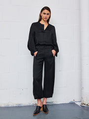 A woman stands against a white brick wall, wearing the Zero + Maria Cornejo Aissa Cargo Pant in black striped jacquard with a matching blouse and black slip-on shoes. Her hands are in her pockets and she has a serious expression.