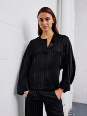 A person with long brown hair stands against a white brick wall, hands in their pockets and looking at the camera, wearing the loose-fitting Long-Sleeved Akeo Shirt by Zero + Maria Cornejo.