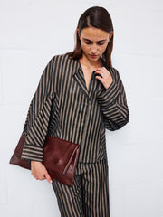 Wearing the Long-Sleeved Akeo Shirt by Zero + Maria Cornejo, a woman holds a large brown leather clutch as she stands against a white brick wall, looking down with her hair parted to the side.