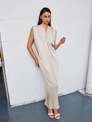 A woman with long brown hair and a calm expression stands against a white brick wall, wearing the Zero + Maria Cornejo Long Issa Dress—a sleeveless, full-length dress made from responsibly-sourced viscose with striped jacquard detailing—paired with sandals.