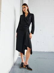 A person stands indoors against a white wall, wearing the Zero + Maria Cornejo Long-Sleeved Madira Dress paired with black shoes. The floor is gray, and the room is minimally decorated.