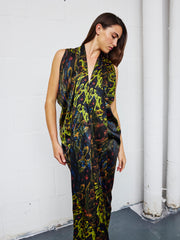 Wearing the Eve Long Foil Dress by Zero + Maria Cornejo, a person stands against a white brick wall with visible pipes, looking down and to the side.