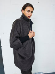 A person with dark hair pulled back wears the Reversible Rio Peacoat by Zero + Maria Cornejo, hands holding the front closed, standing against a white tiled wall.