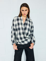 A person with straight brown hair wears the black and white cotton-viscose Jazmin Shirt by Zero + Maria Cornejo paired with black pants, standing against a white tiled wall with their hands resting on their thighs.