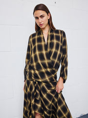 Wearing the Jazmin Shirt by Zero + Maria Cornejo styled as a wrap dress, a person with straight brown hair stands against a white brick wall and looks at the camera with a neutral expression.