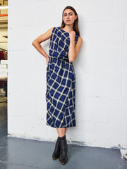 A woman wearing the Elise Dress by Zero + Maria Cornejo—a sleeveless cotton-viscose plaid piece—stands against a white brick wall, one hand on her hip and the other at her side, paired with black ankle boots.