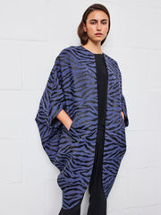 A person stands against a white wall, wearing the Zero + Maria Cornejo Koya Coat—featuring a loose, cocoon silhouette in blue and black tiger stripes—layered over black clothing, with hands tucked into the coat’s pockets.
