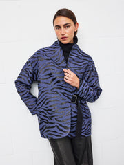 A person stands against a white wall, wearing the Zero + Maria Cornejo Wrap Ari Jacket—a purple and black tiger-motif piece with a stand collar—styled over a black turtleneck and black pants, with one hand holding the jacket lapel.