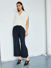 A woman stands indoors against a white brick wall wearing a white V-neck top, the Ruched Front Pant in black by Zero + Maria Cornejo, and black heeled mules—an effortlessly chic look made in New York.