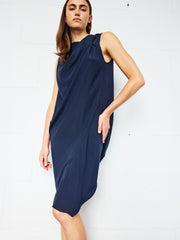 A person with long brown hair wears the Zero + Maria Cornejo Twist Shoulder Ibit Dress—a sleeveless, knee-length navy blue draped dress made in New York—while standing against a white brick wall, gazing confidently at the camera.