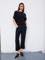 A person stands indoors against a white brick wall, wearing the loose-fitting Eko Pant by Zero + Maria Cornejo—crafted in New York from draped silk—and a matching black top. They have their hands in their pockets, wear black slip-on shoes, and have long dark hair.