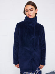 A person wearing the Zero + Maria Cornejo Libi Coat in dark blue wide-wale corduroy, featuring a high collar, stands against a white tiled wall and looks downward with a neutral expression.