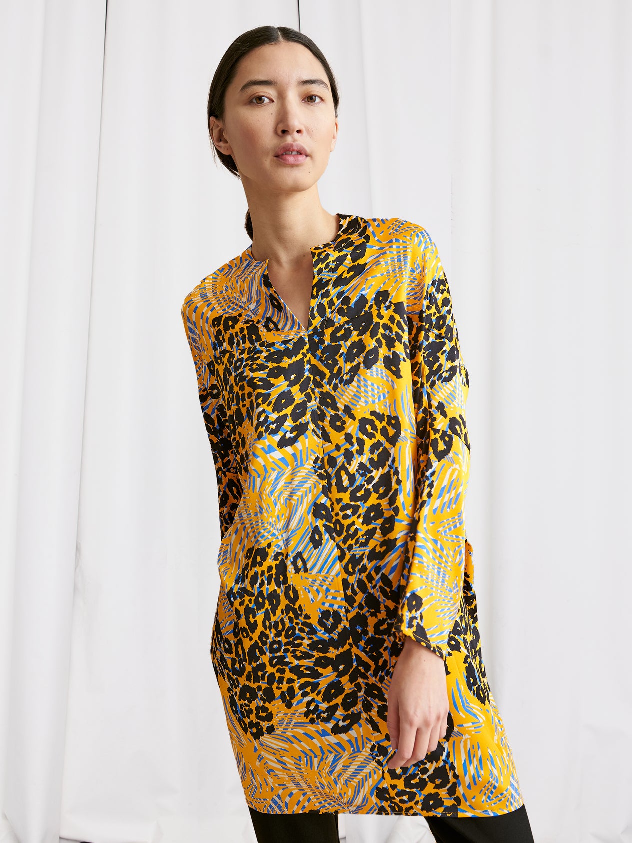 A person stands against a white curtain wearing the Zero + Maria Cornejo Long-Sleeved Hex Tunic Dress, featuring a multicolor animal print with bold black, blue, and orange geometric accents and rolled-up sleeves, posing with relaxed confidence.