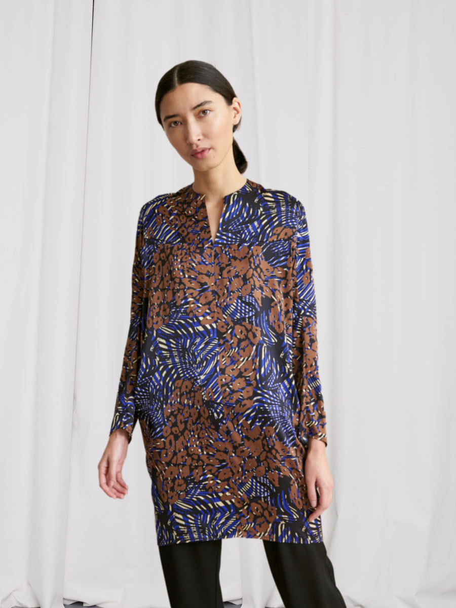 A woman with straight dark hair stands before white curtains, wearing the Zero + Maria Cornejo Long-Sleeved Hex Tunic Dress with a blue and brown abstract print over black pants, her left hand by her side and expression neutral.
