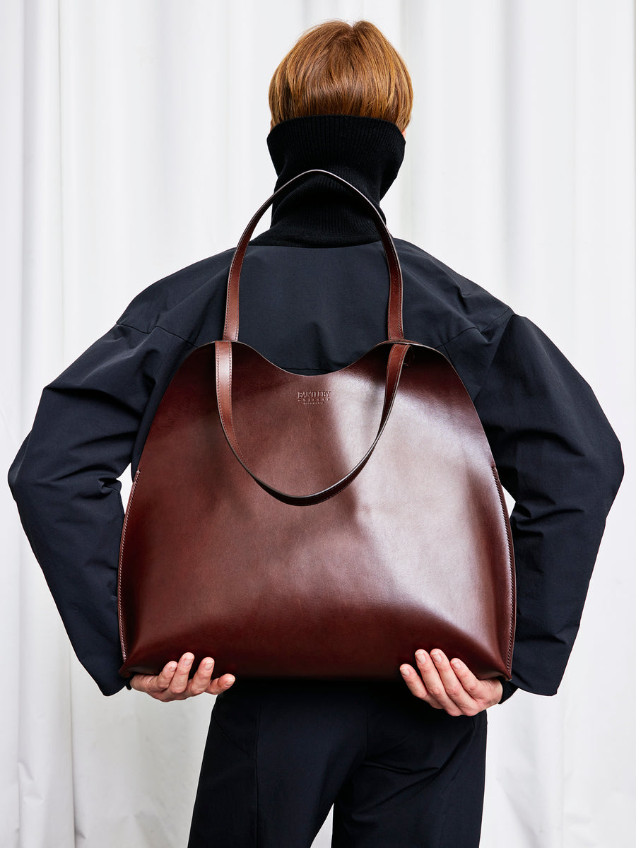 A person in black faces away, holding the chocolate Bartleby Objects Fawkes Tote with two handles. The background is white with soft fabric folds.