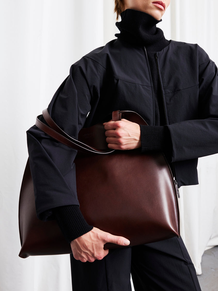 A person in a black long-sleeve jacket holds the large, dark brown Bartleby Objects Fawkes Tote under their arm against a white background. Their face is partially out of frame.