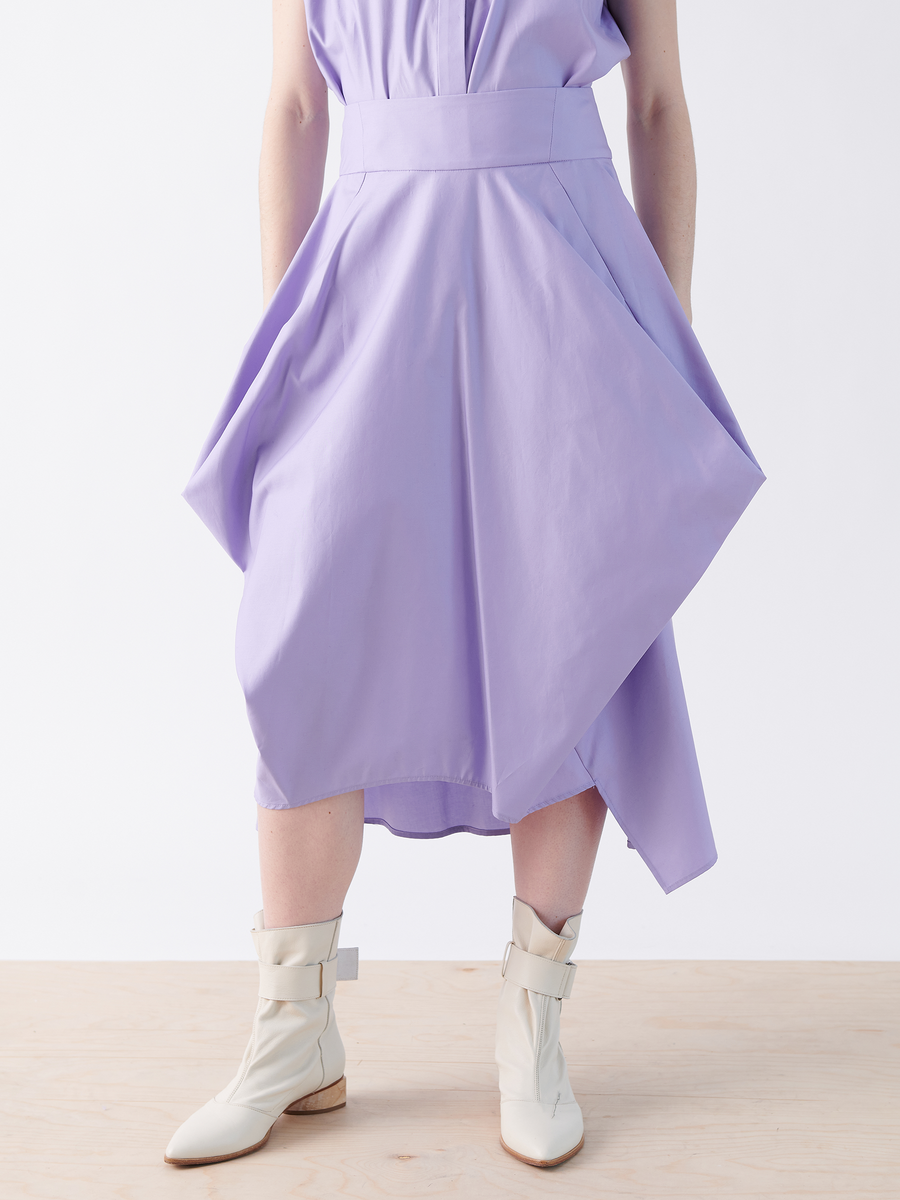 Someone wears a Nio Skirt by Zero + Maria Cornejo in light purple with an asymmetrical design and flared silhouette. They pair it with white ankle boots featuring buckles, standing elegantly on a light wooden floor against a crisp white backdrop.