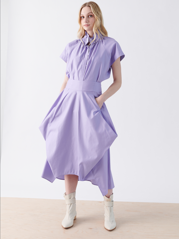 A woman models the Nio Skirt by Zero + Maria Cornejo, a light purple dress with short sleeves, a seamed waist, and full skirt. Styled with white ankle boots, her hands rest in the pockets. The geometric silhouette contrasts beautifully against a plain white background on a light wooden floor.