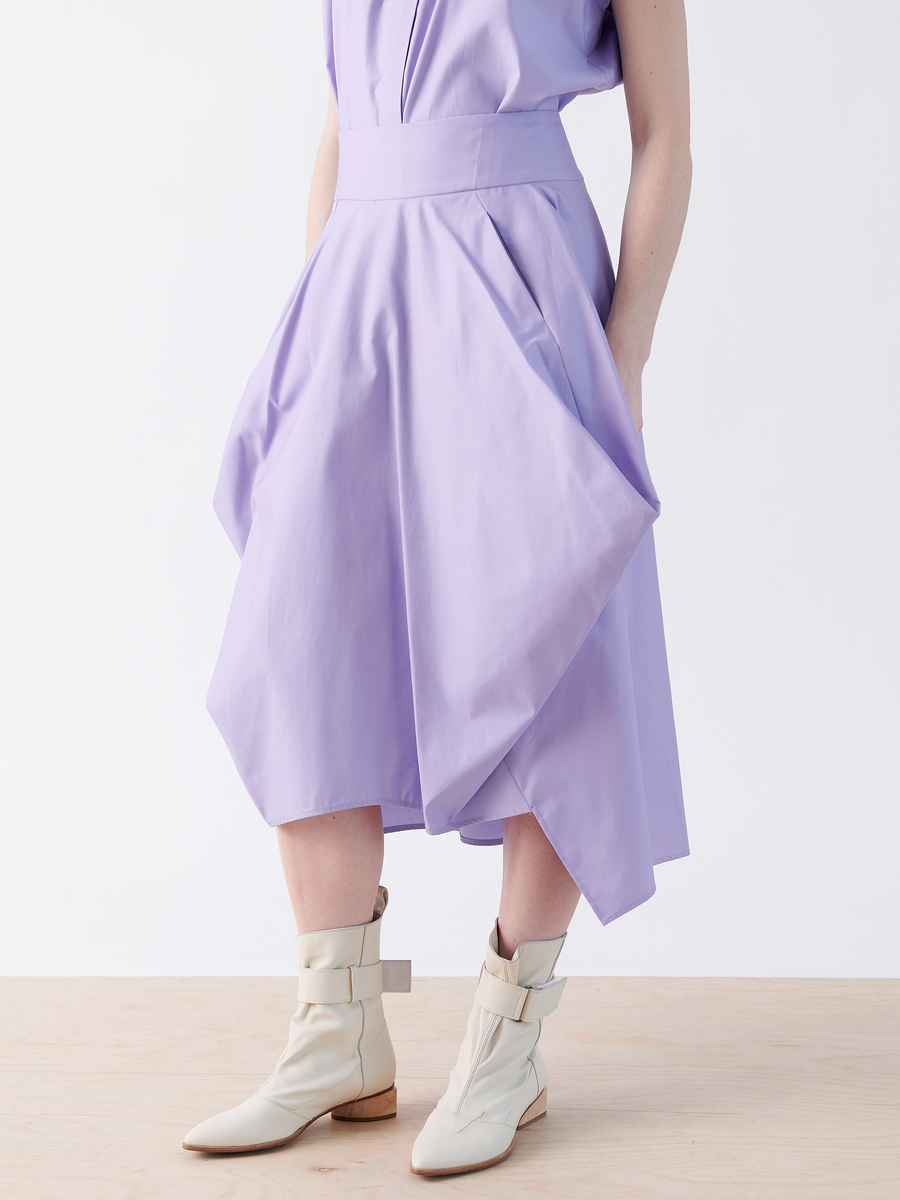 A person wearing Zero + Maria Cornejo's Nio Skirt in lavender—a cotton asymmetrical design with a seamed waist—paired with beige ankle boots featuring straps, stands against a plain white background.