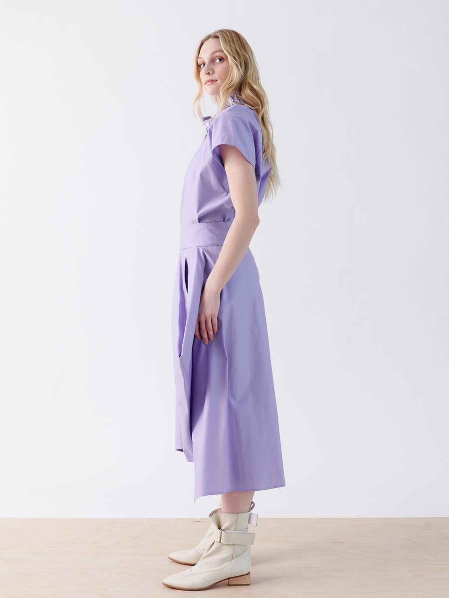 A person with long blonde hair wears the Nio Skirt by Zero + Maria Cornejo—light purple with a seamed waist—and cream-colored boots, standing sideways, gazing over their shoulder. The background is plain white.