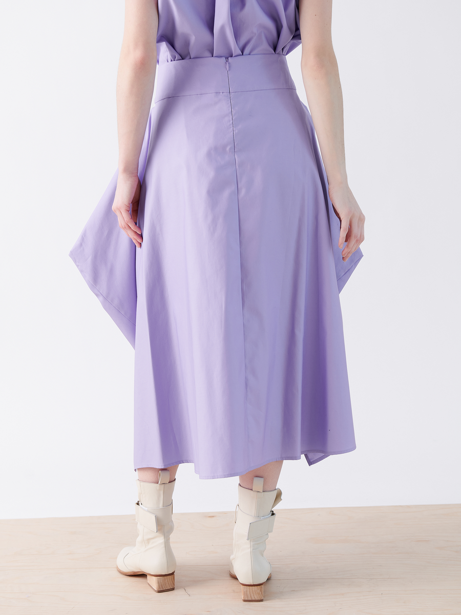 A person wearing the Nio Skirt - Final Sale by Zero + Maria Cornejo, a light purple skirt with a geometric silhouette and asymmetrical hem, pairs it with a top and cream ankle boots. The scene features a white background and light wood flooring.