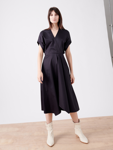 On a light wood floor, someone wears the Aki Wave Dress by Zero + Maria Cornejo, featuring a dark, knee-length wrap-effect with short sleeves and a V-neckline. Its asymmetrical skirt is paired with off-white ankle boots against a brightly lit background that enhances the outfit's elegance.