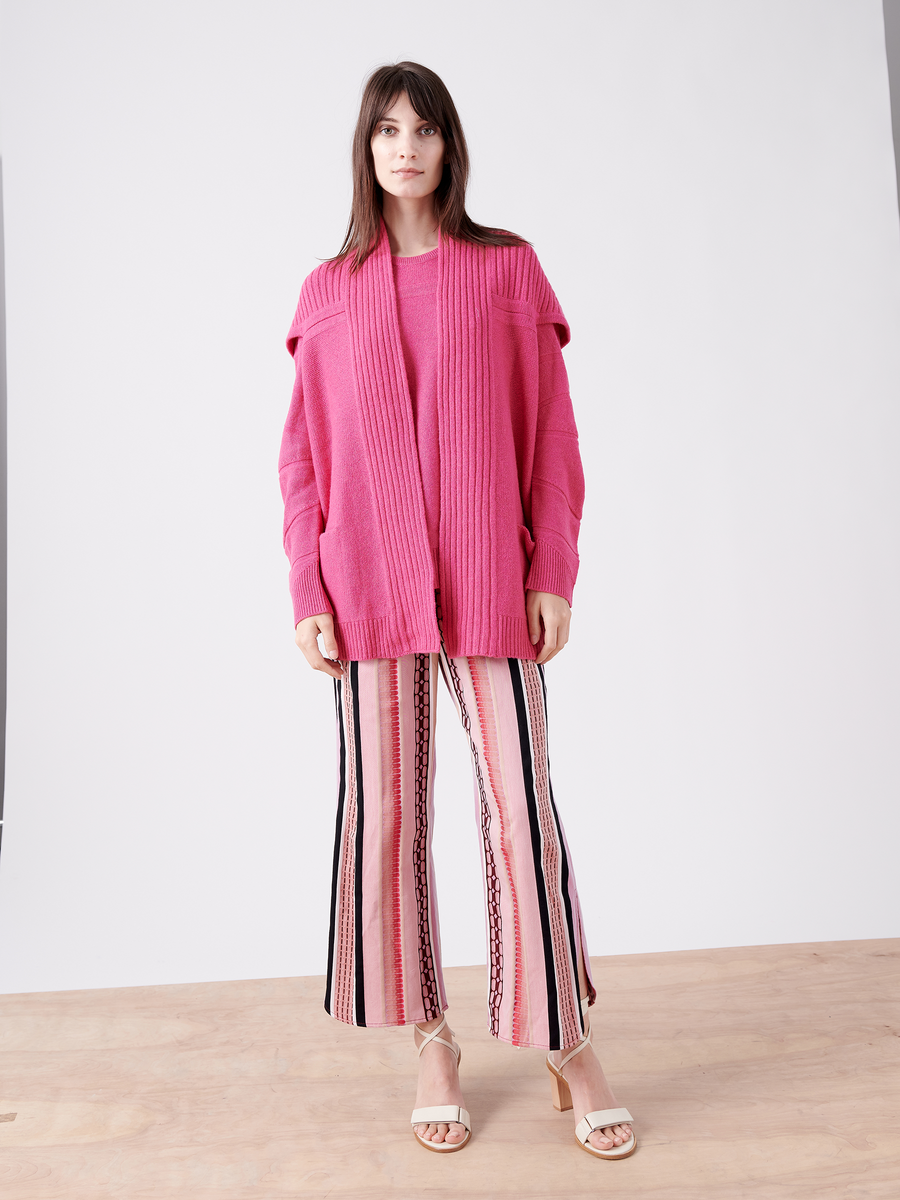 A woman stands on a wooden floor wearing the eco-friendly Nian Shrug by Zero + Maria Cornejo over a matching top, paired with vibrant pink striped wide-leg pants. Her long, straight hair complements her white open-toed heels against a plain white wall backdrop.