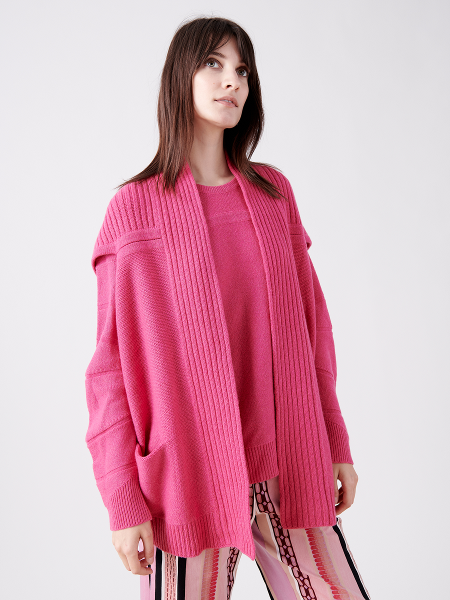 A woman in a pink cardigan, the Nian Shrug by Zero + Maria Cornejo, crafted from eco-friendly yarn, stands against a white background. Her light patterned pants complement her sustainable fashion choice. With long brown hair, she gazes slightly upwards, embracing style with conscience.