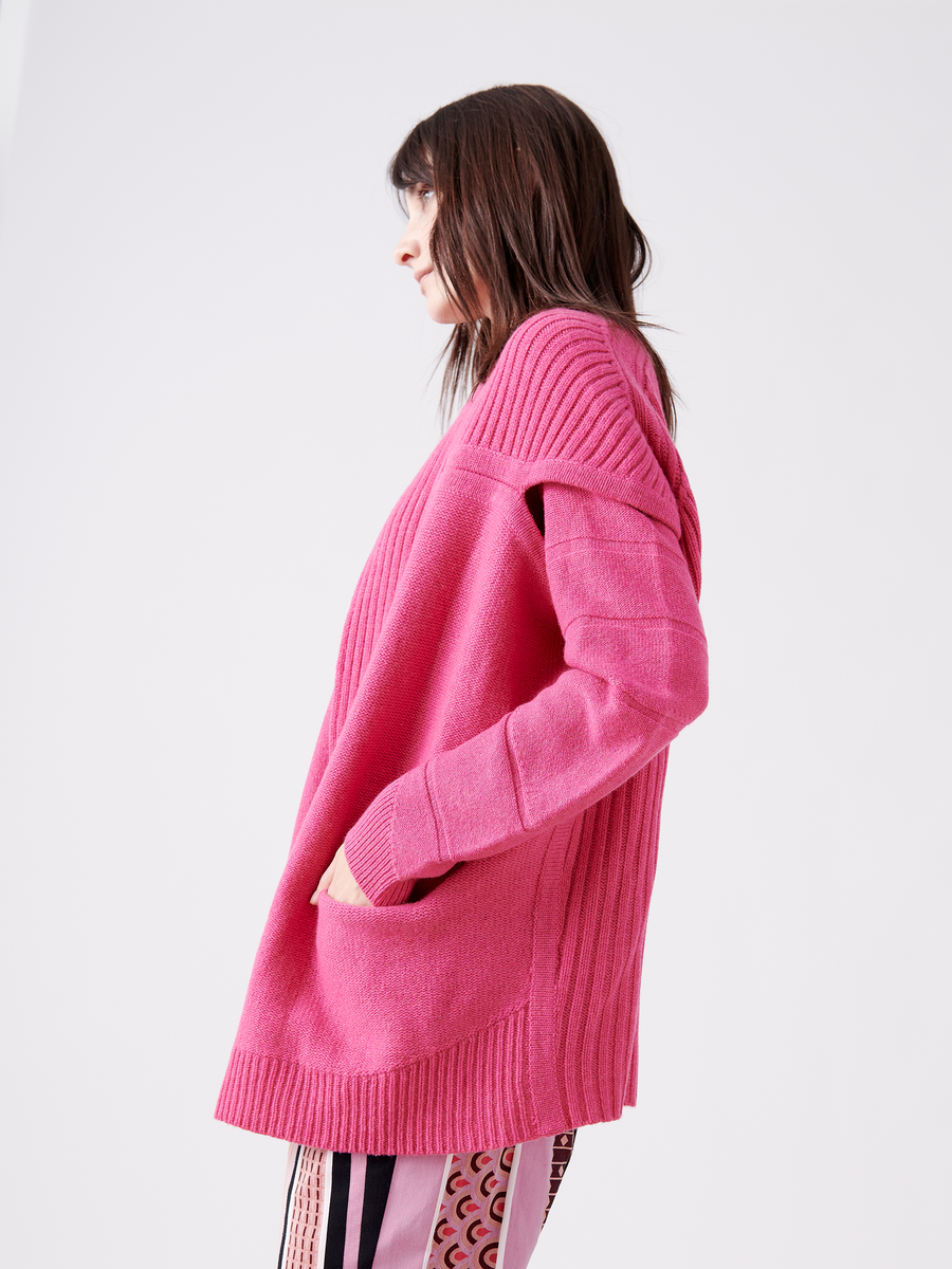 A person with long hair wears the eco-friendly Nian Shrug in pink by Zero + Maria Cornejo, paired with patterned pants. They stand sideways, hand in pocket, against a white background, embodying sustainable fashion.