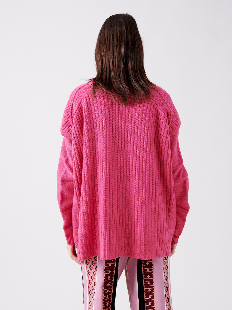 A person with long brown hair is wearing the eco-friendly Nian Shrug, an oversized ribbed sweater in pink by Zero + Maria Cornejo, and patterned pants with vertical burgundy stripes. They face away from the camera against a plain white backdrop, emphasizing their sustainable fashion choice.