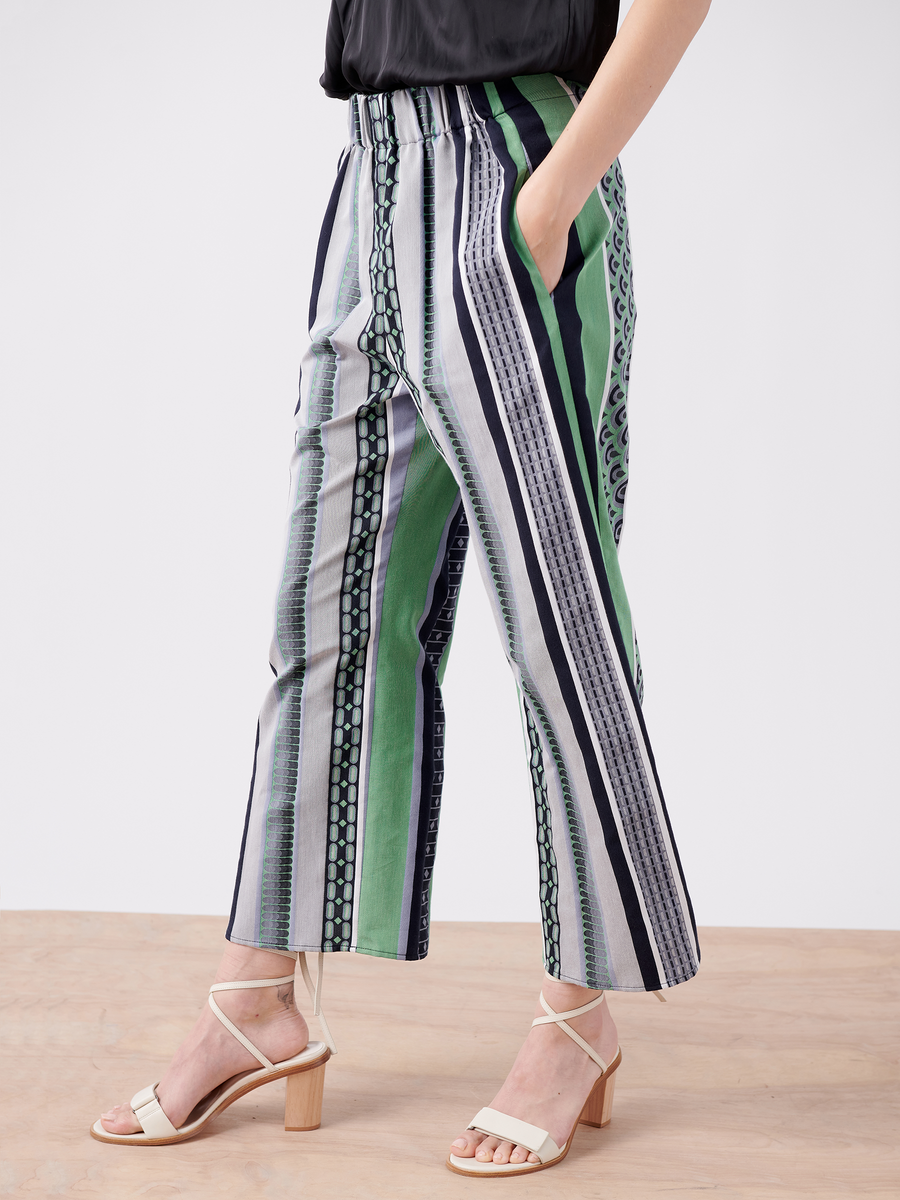 Wearing Zero + Maria Cornejo's Ruched Front Pant - Final Sale in green, black, white, and blue stripes with a black top, they stand on a wooden floor with their right hand in a pocket and complement the outfit with white high-heeled sandals.