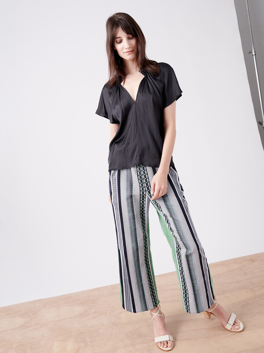 In a studio setting, a woman wears Zero + Maria Cornejo's Ruched Front Pant—GOTS-certified striped jacquard cropped pants in white, black, and green—with a loose black blouse and white sandals. Her hair falls naturally past her shoulders against a plain light gray backdrop.