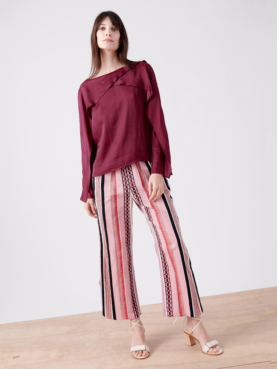 Standing on a light wooden surface, an individual wears Zero + Maria Cornejo's Ruched Front Pant - Final Sale. The burgundy long-sleeve blouse's angled design complements the pink striped jacquard cropped pants made from organic cotton. Their long hair and light-colored heels appear against a plain white background.