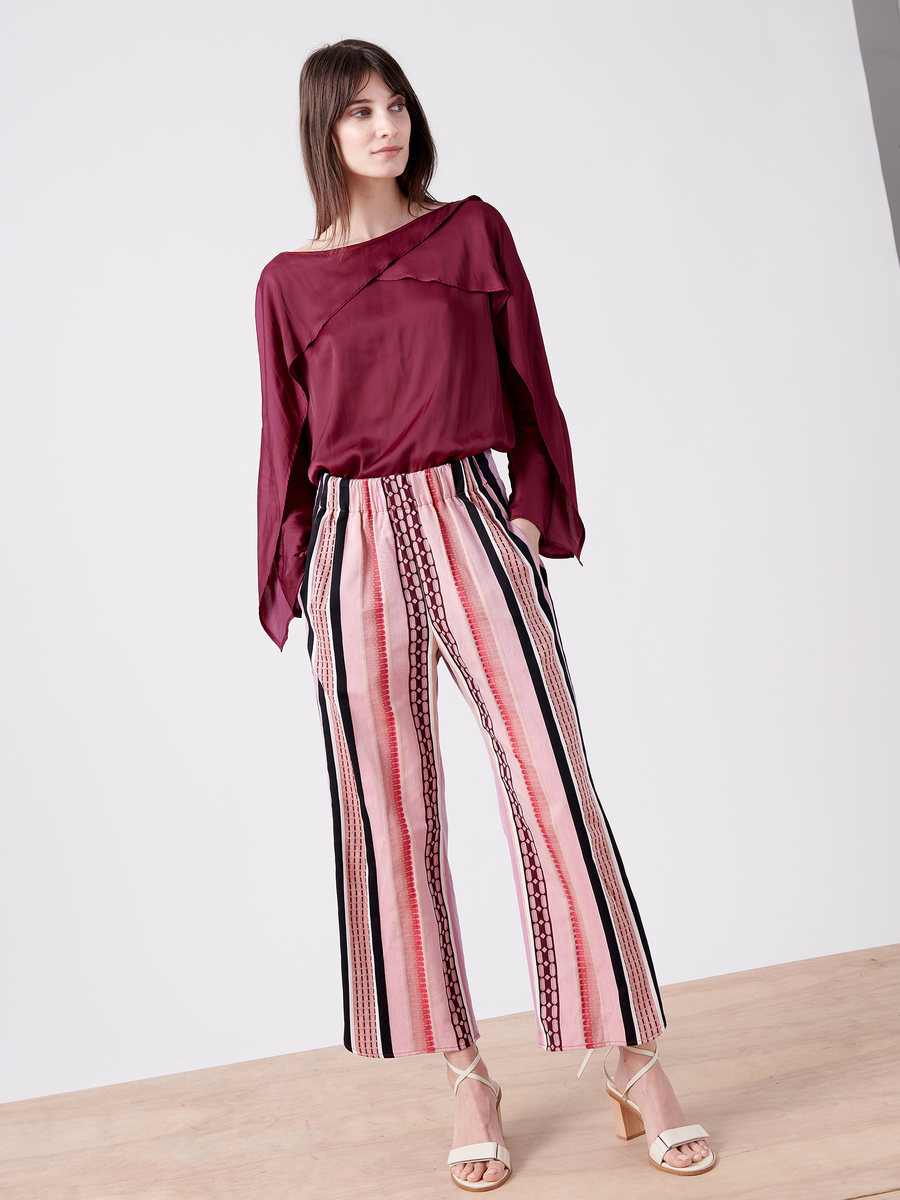 A person stands confidently in a burgundy long-sleeve top and Zero + Maria Cornejo Ruched Front Pant with pink, black, and white stripes on organic cotton. They stand on a light wood floor against a plain background, wearing GOTS certified strappy heeled sandals.