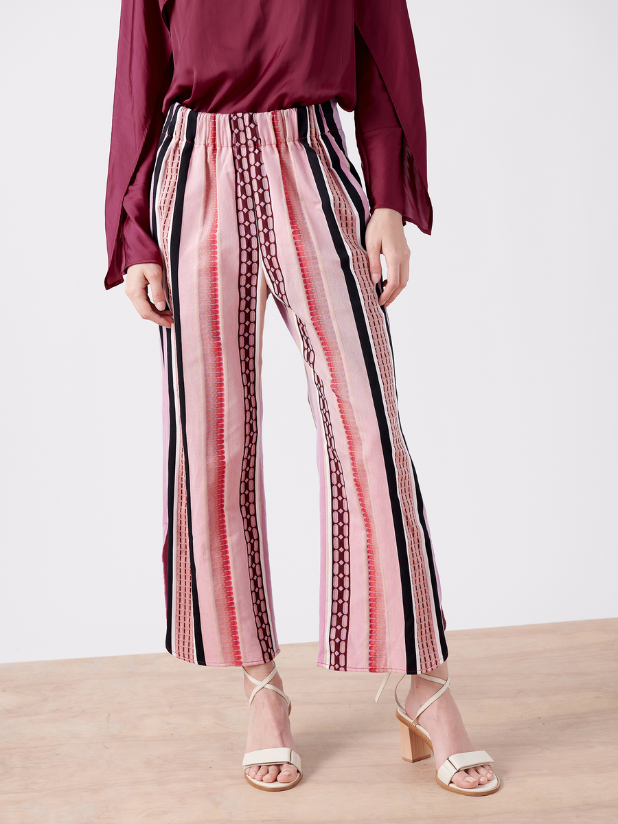 A person wears Zero + Maria Cornejo's Ruched Front Pant, a GOTS certified striped jacquard cropped pant with vertical red, pink, black, and white stripes. Paired with a burgundy long-sleeved blouse and strappy white heels, they stand on a wooden floor against a plain background.