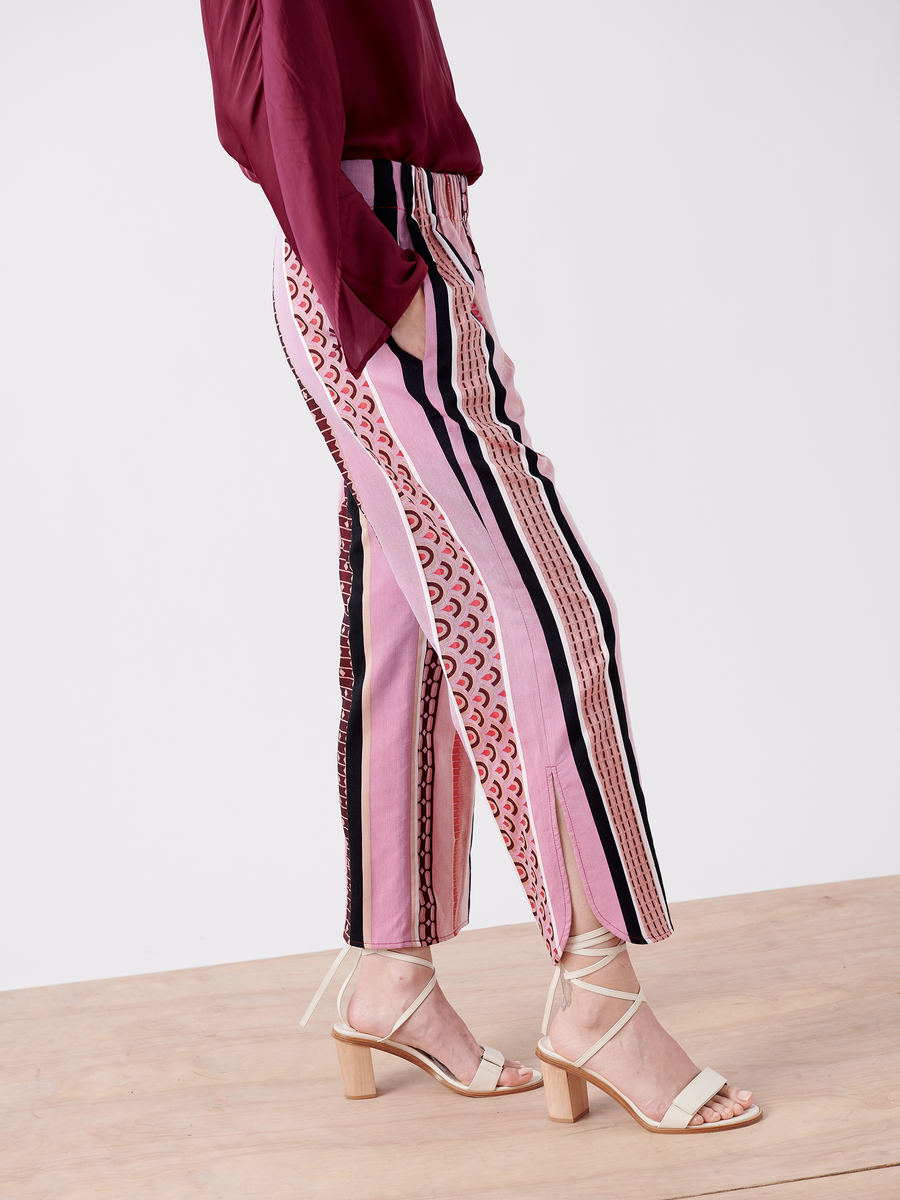 A person in the Ruched Front Pant - Final Sale by Zero + Maria Cornejo, featuring striped jacquard in pink and black GOTS certified organic cotton, paired with a maroon top. They stand on a wooden floor wearing ankle-strap block-heeled sandals against a plain white backdrop.