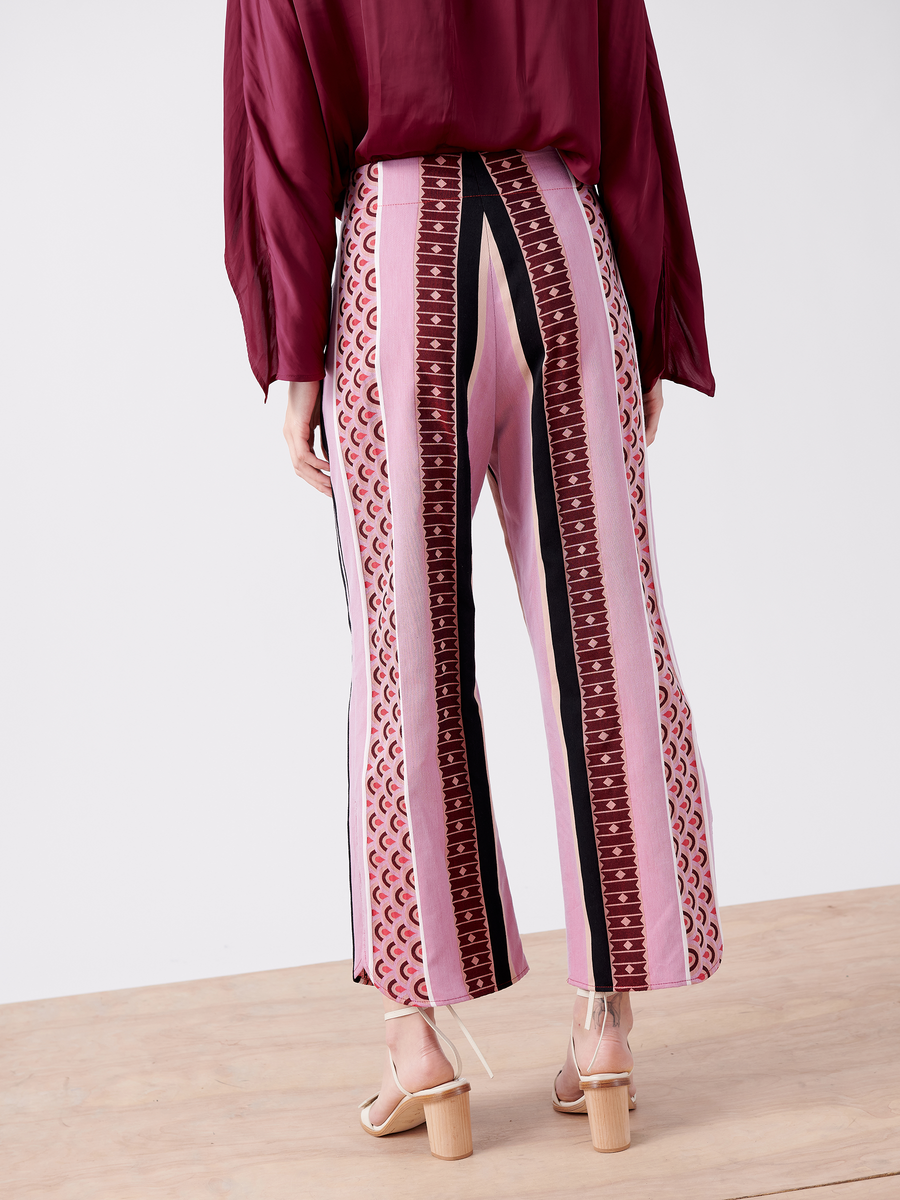 A person is seen from behind wearing Ruched Front Pants by Zero + Maria Cornejo. The high-waisted, wide-leg design features vertical pink, black, and maroon geometric patterns in organic cotton. They pair the pants with a long-sleeve maroon top and beige open-toe block heels on a wooden floor.