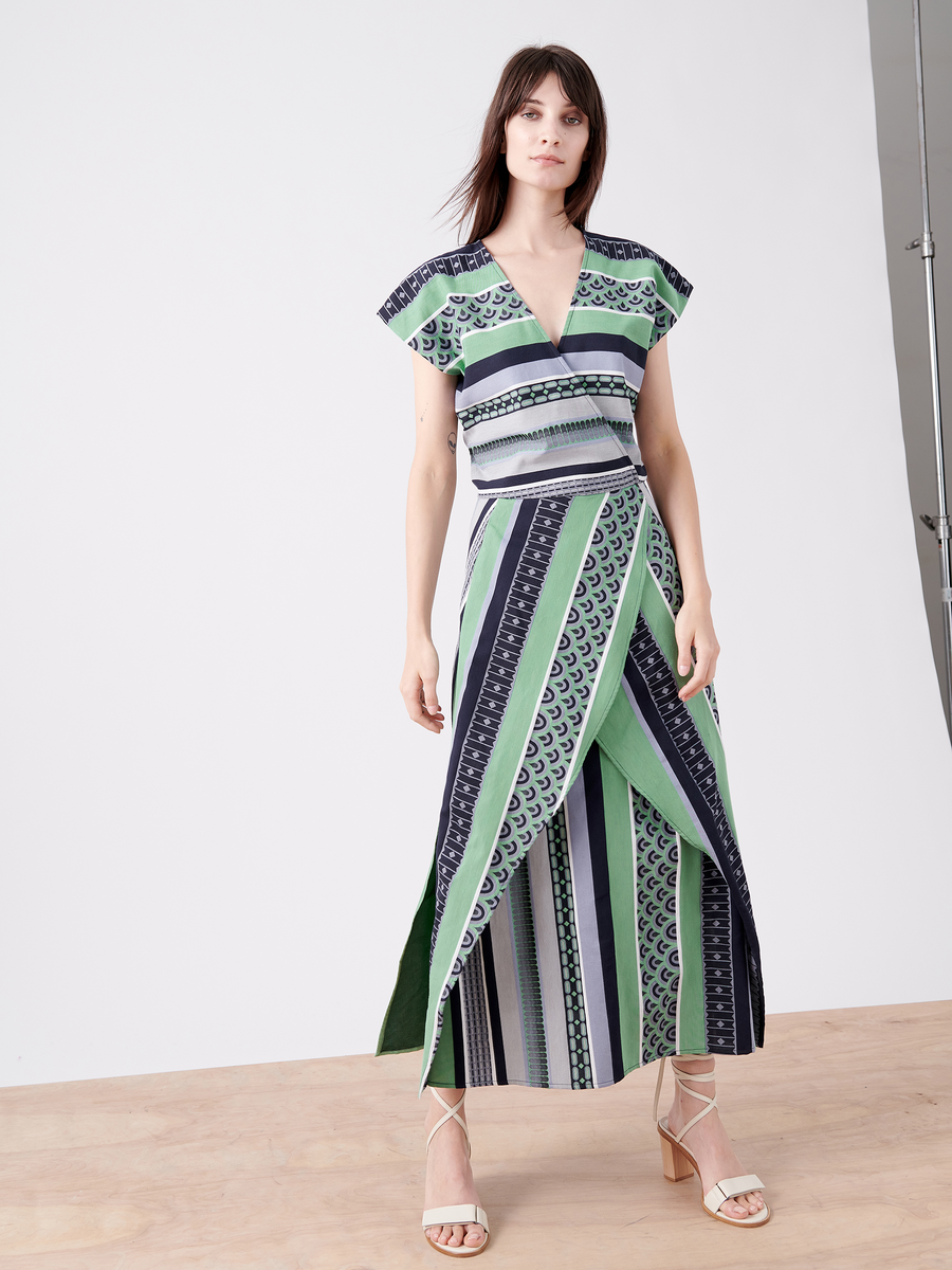 A woman poses elegantly on a light wooden floor, donning the Mido Dress by Zero + Maria Cornejo, which features green, blue, and white striped jacquard patterns. The wrap-effect V-neck enhances its abstract design. She steps confidently in nude heels, her long dark hair flowing against a white background.