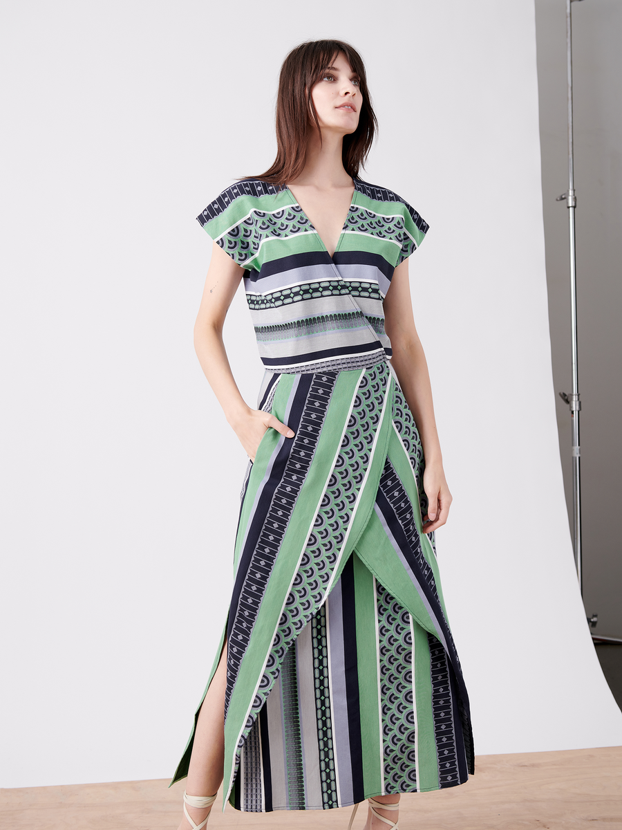 A person in the Mido Dress by Zero + Maria Cornejo poses against a plain backdrop. The green, black, and white striped jacquard dress, made from organic cotton, features short sleeves, a wrap-effect V-neck, and a long skirt with a front slit. The individual stands on a wooden floor.